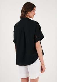 Black short-sleeve shirt featuring eyelet detailing on sleeves, a relaxed fit, and a rounded hem. Paired with white shorts.