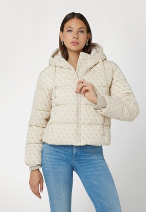 Vrouw in een beige, geblokte pufferjack met capuchon, tot de helft dichtgeritst, met lichtblauwe jeans, poserend tegen een effen lichte achtergrond.