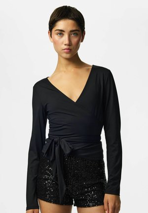 Jeune femme aux cheveux courts et foncés portant un haut cache-cœur noir à manches longues et un short noir à sequins, debout devant un fond blanc.