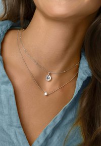 Tre sølvhalskæder lagdelt omkring en hals: et med en cirkulær vedhæng med en indgraveret "M," et andet med små perler, og et med en perle.