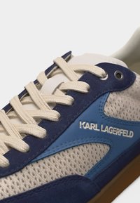 Κοντινή λήψη μπεζ και μπλε αθλητικού παπουτσιού με εκρού κορδόνια, ύφασμα από δίχτυ, λεπτομέρειες από σουέντ και λογότυπο "KARL LAGERFELD" στο πλάι.