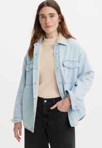 Lys blå denimjakke med en myk beige pelshals, to brystlommer og knappestang. Brukes over en beige topp og svarte jeans.
