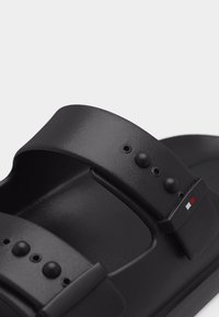 Μαύρο αναπληρώσιμο σανδάλι με δύο λουριά που διαθέτουν στρογγυλά κουμπιά και μικρή ταμπέλα με λογότυπο σε κόκκινο, άσπρο και μπλε χρώμα.