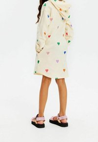 Persona che indossa un vestito a cappuccio color crema con stampe di cuori multicolori e sandali platform rosa, in piedi con le mani nelle tasche.