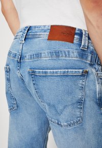 Lichtblauwe denim jeans met een gestructureerde afwerking, voorzien van een bruine leren patch op de tailleband en kenmerkende stiksels op de achterzakken.