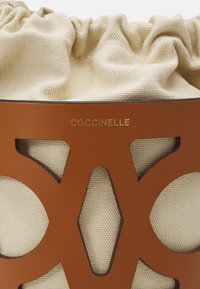 Tan leather bag with cut-out designs and a cream drawstring top, featuring the embossed brand name "Coccinelle" in gold.