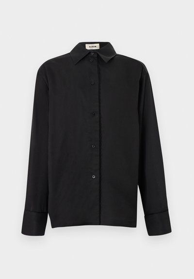 Μαύρο πουκάμισο με μακριά μανίκια και κουμπιά, με μυτερό γιακά και μανσέτες, απλωμένο επίπεδα με την ετικέτα της μάρκας "Aeron" ορατή μέσα στον γιακά.