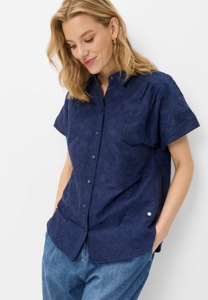 Femme aux cheveux blonds portant une chemise bleu marine à manches courtes avec une broderie florale subtile et un jean bleu clair, les mains dans les poches.