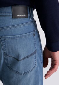 Die Gesäßtasche von hellblauen Jeans mit einem schwarzen Lederpatch, auf dem "pierre cardin" steht, und einer Hand neben der Hüfte.