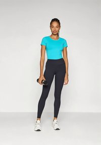 Camiseta ajustada de manga corta color turquesa, leggings negros de cintura alta y zapatillas deportivas blancas. La persona sostiene una mancuerna negra en una mano.