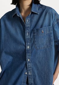 Dunkelblaue Jeanshemd mit Knopfverschluss, spitzem Kragen, Brusttasche und kleinem aufgesticktem Logo auf der Tasche.