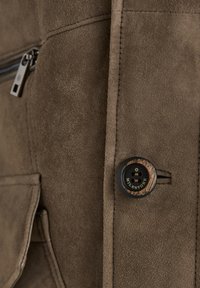 Nahaufnahme einer braunen Wildlederjacke mit einem schwarzen Knopf mit holzähnlichen Akzenten und einer Reißverschlusstasche, die eine glatte Textur zeigt.