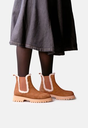 Bottes marron à la cheville avec bordure en fourrure beige portées avec des collants noirs et une jupe foncée longueur genou sur fond blanc.