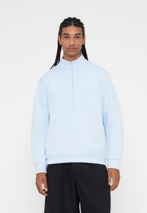 Sweat-shirt bleu clair à zip quart avec une texture douce, des poignets côtelés et un design en panneaux. Associé à un pantalon noir ample.