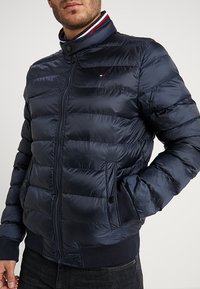 Granatowa kurtka puchowa z pikowanym wzorem, wysoki kołnierz, zamek błyskawiczny, kieszenie boczne i ściągacze przy mankietach. Zawiera logo na piersi i kontrastowe wykończenie.