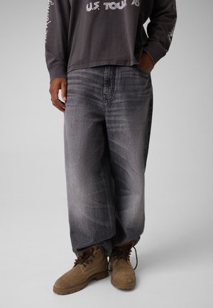 Modèle portant un jean ample gris anthracite, un sweat-shirt noir à motif graphique et des bottes marron à lacets, sur un fond de studio uni.