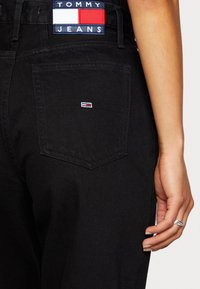 Lähikuvana mustat Tommy Jeans farkut, joissa on merkkilogo takataskun yläpuolella ja pieni lippulogo taskussa, käsi lepää farkkujen vieressä.