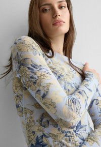 Joven con cabello largo y castaño, vistiendo una blusa de manga larga de color azul claro con patrones florales beige y azules, posando frente a un fondo liso.