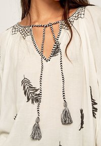 Leichte, weiße Bluse mit schwarzer Blätterstickerei und einem V-Ausschnitt, dessen Kordel gestreift und zum Binden ist. Dekorative Quasten hängen von der Kordel.