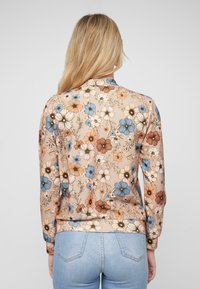 Chemise à motifs floraux en beige, ornée de fleurs bleues, brunes et crème. Fabriquée en tissu léger, elle possède des manches longues et un col rond.