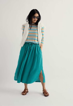 Kvinde iført solbriller, stribet langærmet skjorte, hvid sweater draperet over skuldrene, grønblå nederdel med sidelomme og brune sandaler, stående.