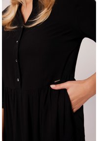 Czarna sukienka z guzikami z przodu i krótkimi rękawami, z marszczonym pasem oraz dyskretną kieszonką z boku. Miękka tekstura materiału.