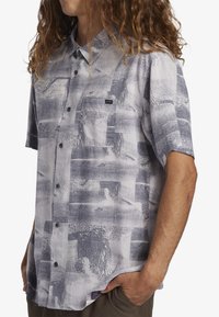 Camicia a maniche corte di colore grigio chiaro con un motivo astratto nero a trama. Presenta una chiusura frontale con bottoni e una tasca sul petto.