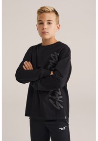 Schwarzes Langarm-T-Shirt aus Baumwolle mit einem großen grauen Grafikdruck auf der Vorderseite und gekrempelten Ärmeln. Kombiniert mit schwarzen Hosen.