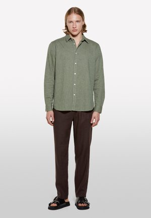 Chemise verte à boutons avec un tissu texturé, associée à un pantalon marron et des sandales noires. La chemise possède un col et une fermeture à boutons.