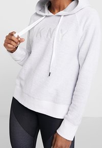 Ljust grå hoodie i texturerad tyg, vita dragsko, och en diskret präglad "DKNY"-logotyp på framsidan. Vita manschetter.
