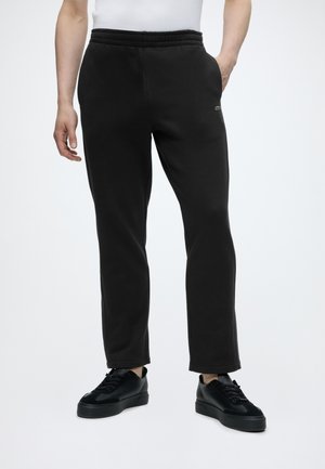 Mann trägt schwarze eng anliegende Sweatpants mit Seitentaschen und schwarze Sneakers, steht vor einem einfarbigen Hintergrund.