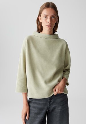 Giovane donna con capelli castano chiaro che indossa un maglione a coste verde chiaro con un ampio colletto e jeans grigio scuro, in posa con una mano in tasca.