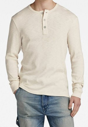 Mann trägt ein beiges Langarm-Henley-Shirt mit drei Knöpfen und hellblaue Jeans, die Hände entspannt an den Seiten.