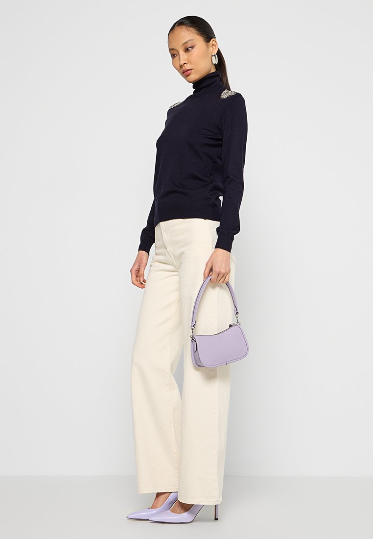 Donna che indossa un dolcevita nero, pantaloni larghi colore crema, tacchi lilla e tiene in mano una piccola borsa lilla, in piedi su uno sfondo neutro.