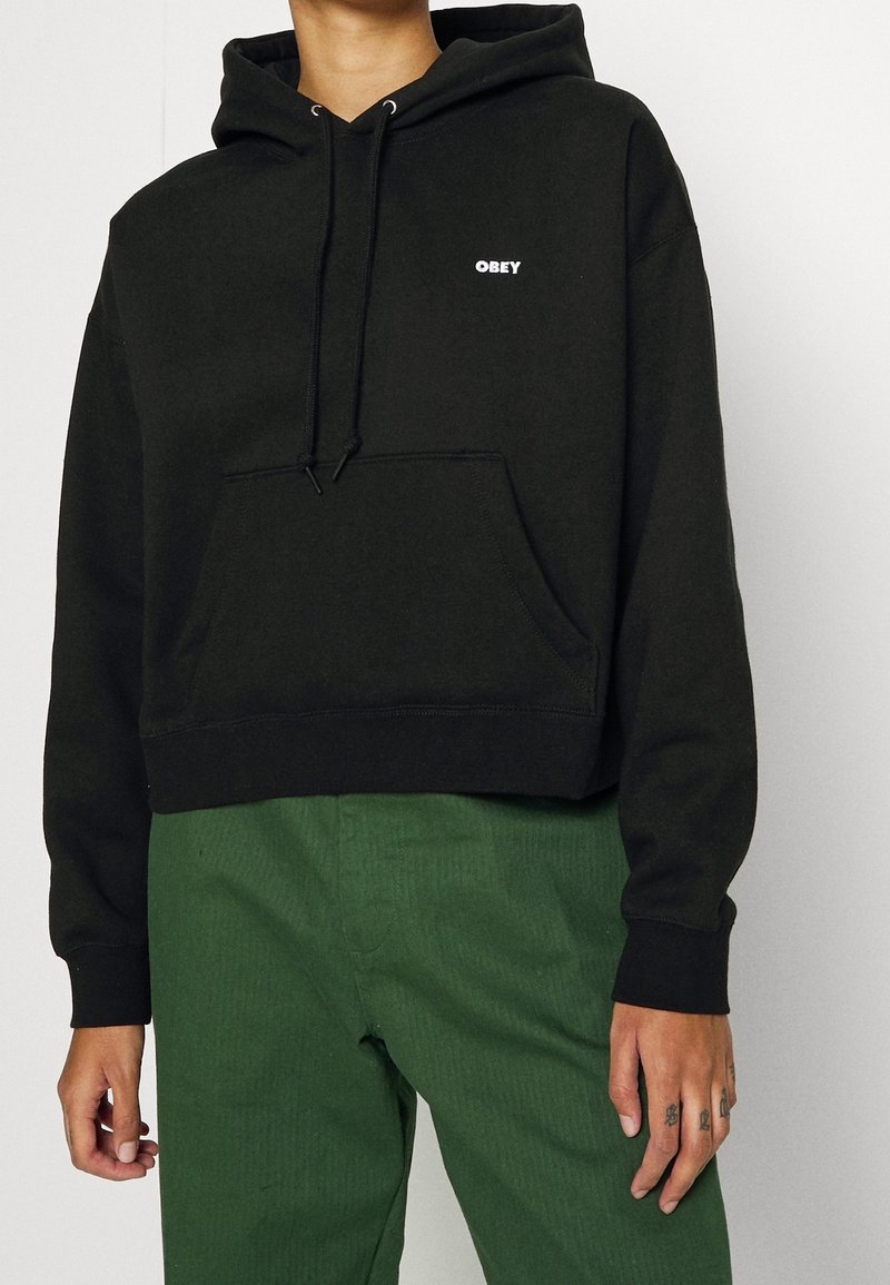 Černý zkrácený mikina s kapucí z měkké látky, s bílým logem "OBEY" na hrudi, přední kapsou a nastavitelnou šňůrkou na kapuci.