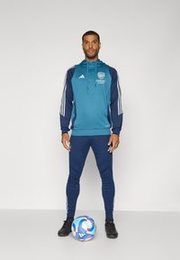 Blue hooded sweatshirt with navy accents, white Adidas stripes, and Arsenal branding. Fitted navy leggings and black soccer shoes. Soccer ball at feet.