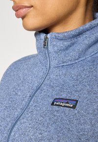 Giacca in pile con zip, di un blu texturizzato, con colletto alto e cerniera argentata. Presenta un logo Patagonia nero sul petto.