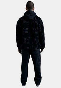 Chaqueta holgada de estampado negro con acentos morados, llevada sobre un sudadera con capucha oscura y pantalones cargo negros. Un aspecto general de capas casual.