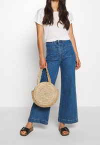 Modré džíny s vysokým pasem a širokými nohavicemi, se dvěma zadními kapsami, v kombinaci s bílým topem se krajkovými detaily a černými skluznými sandály.