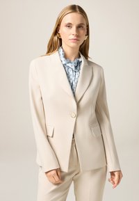 Blazer beige su misura con rever a punta, chiusura con un solo bottone, due tasche anteriori e tessuto liscio. Indossato sopra una blusa blu a fantasia.