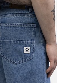Pantaloni scurți din denim de culoare albastru deschis, cu o textură subtilă. Au un buzunar la spate cu un etichetă albă și cusătura logo-ului negru.