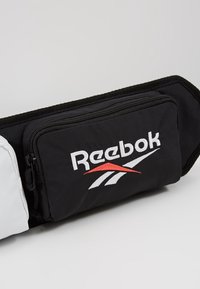 Banane noire Reebok avec logo blanc et rouge et poche principale zippée sur un fond clair uni.