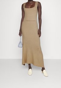 Rippstrick-Maxikleid in Beige mit quadratischem Ausschnitt und figurbetonter Silhouette, kombiniert mit weißen Slippern und einer kleinen blauen Handtasche.