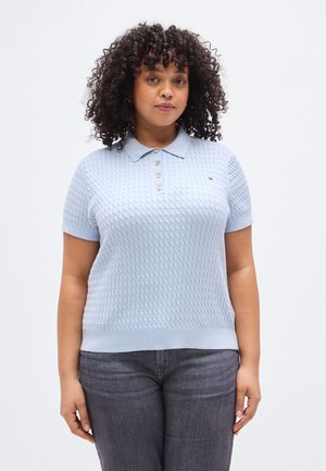 Light blue knitted polo shirt with a textured pattern, short sleeves, and three gold buttons at the collar. Paired with dark gray jeans.