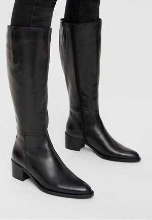 Bottes en cuir noir jusqu'au genou avec un bout pointu et un talon carré. Présentent une fermeture éclair sur le côté et une texture lisse et brillante.