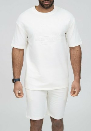 Homme portant un t-shirt blanc à manches courtes et un short blanc assorti, debout les bras détendus, devant un fond clair uni.