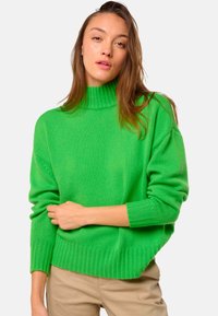 Femme aux longs cheveux bruns portant un pull à col roulé vert vif et un pantalon beige, posant avec un bras croisé sur la taille.