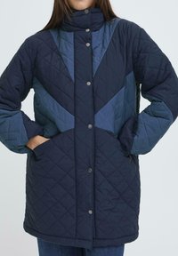 Chaqueta acolchada en azul oscuro con acentos en azul más claro, que cuenta con un alto cuello, botones a presión y bolsillos laterales. Tela texturizada y patrones de diamante.