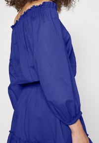 Off-shoulder klänning i livfull blått tyg, med volangkanter, lösa ärmar och samlad midja, mjuk textur med en lätt lyster.