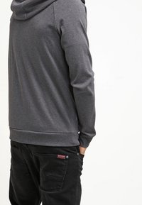 Sweat à capuche gris foncé avec une capuche ample, des poignets côtelés et une coupe décontractée, associé à un pantalon noir avec poches latérales et détail logo.